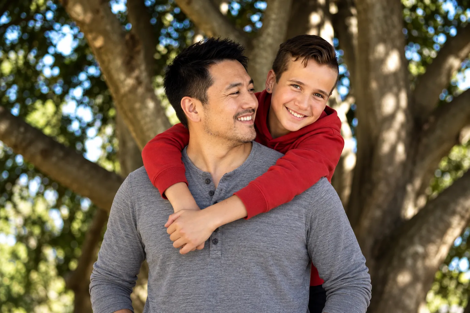 A father carrying his son on his back under a big tree, both smiling