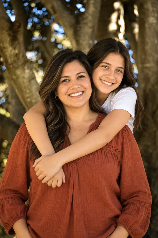 A smiling mother hugged from behind by her teenage daughter under a tree